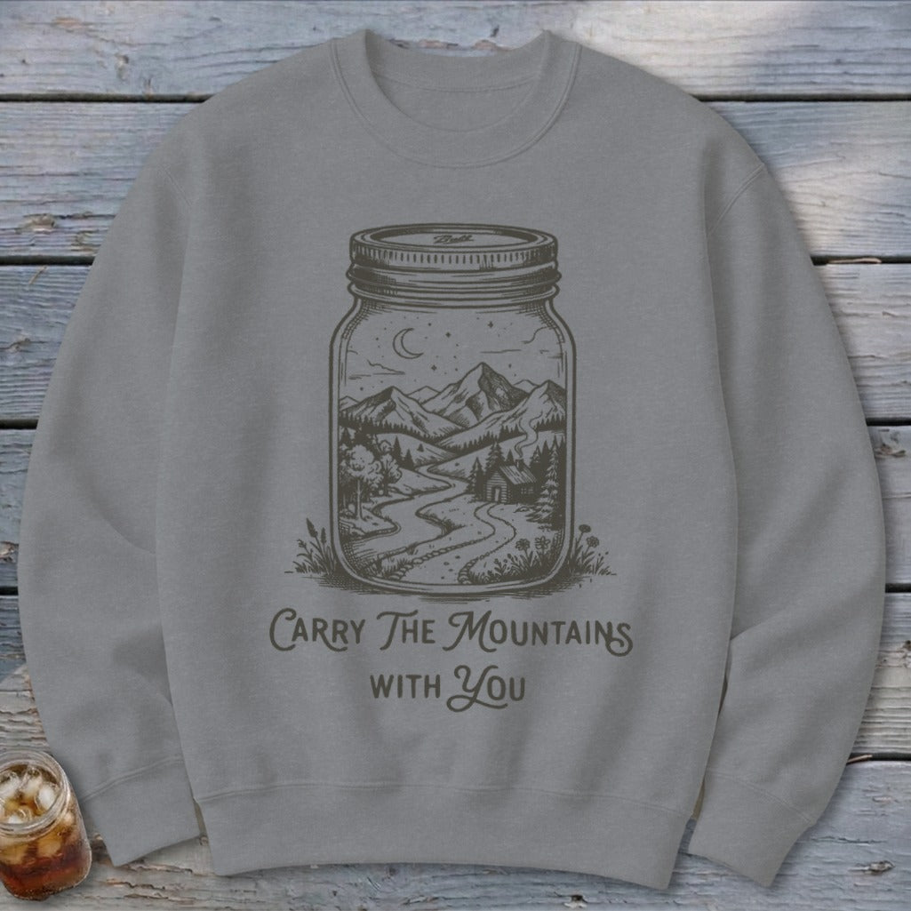 A grey crewneck sweatshirt featuring a graphic of a mountain landscape inside a glass jar, with text reading "Carry the Mountains with You."