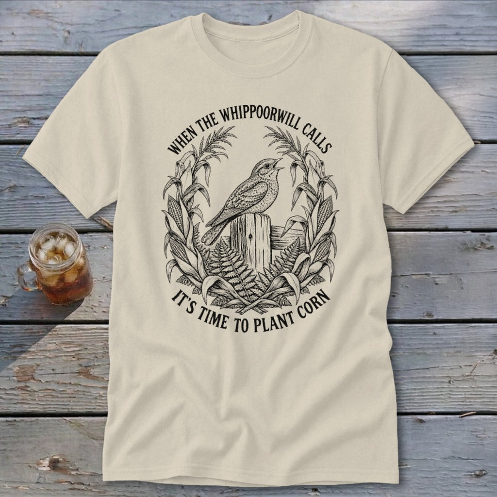 A cream-colored t-shirt with a black line art graphic of a whippoorwill perched on a wooden post, surrounded by corn stalks and ferns, and text that reads "WHEN THE WHIPPOORWILL CALLS IT'S TIME TO PLANT CORN".
