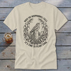 A cream-colored t-shirt with a black line art graphic of a whippoorwill perched on a wooden post, surrounded by corn stalks and ferns, and text that reads "WHEN THE WHIPPOORWILL CALLS IT'S TIME TO PLANT CORN".