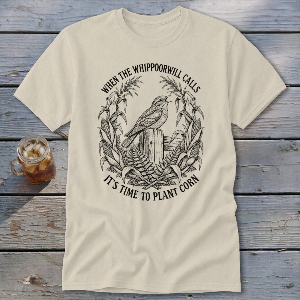 A cream-colored t-shirt with a black line art graphic of a whippoorwill perched on a wooden post, surrounded by corn stalks and ferns, and text that reads "WHEN THE WHIPPOORWILL CALLS IT'S TIME TO PLANT CORN".