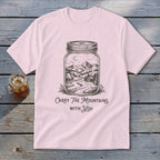 A pink short-sleeve t-shirt with a graphic of mountains and a cabin inside a mason jar and text reading Carry The Mountains With You.