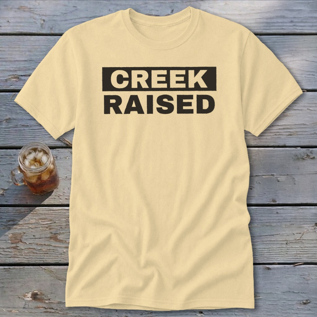 Cream t-shirt with "CREEK RAISED" text in black on a rustic wooden surface.