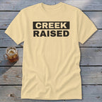 Cream t-shirt with "CREEK RAISED" text in black on a rustic wooden surface.