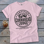 Light pink short-sleeved t-shirt with a round emblem reading "Certified Cornbread Judge" and a glass of iced tea nearby.