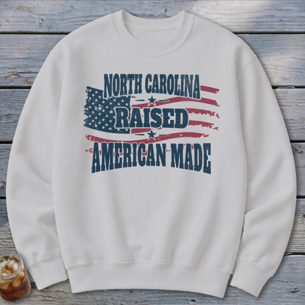Off-white crewneck sweatshirt featuring a distressed American flag graphic with text reading North Carolina Raised American Made.