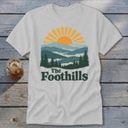 White t-shirt with sunset mountain range graphic and "The Foothills" text.