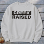 White crewneck sweatshirt with black text "CREEK RAISED"