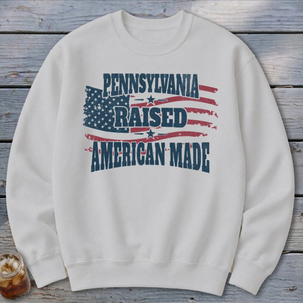 A white crewneck sweatshirt featuring a distressed American flag graphic with the text Pennsylvania Raised American Made.