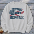 A white crewneck sweatshirt featuring a distressed American flag graphic with the text Pennsylvania Raised American Made.