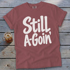 A dusty rose t-shirt featuring the white text Still A Goin lying flat on weathered wooden boards.