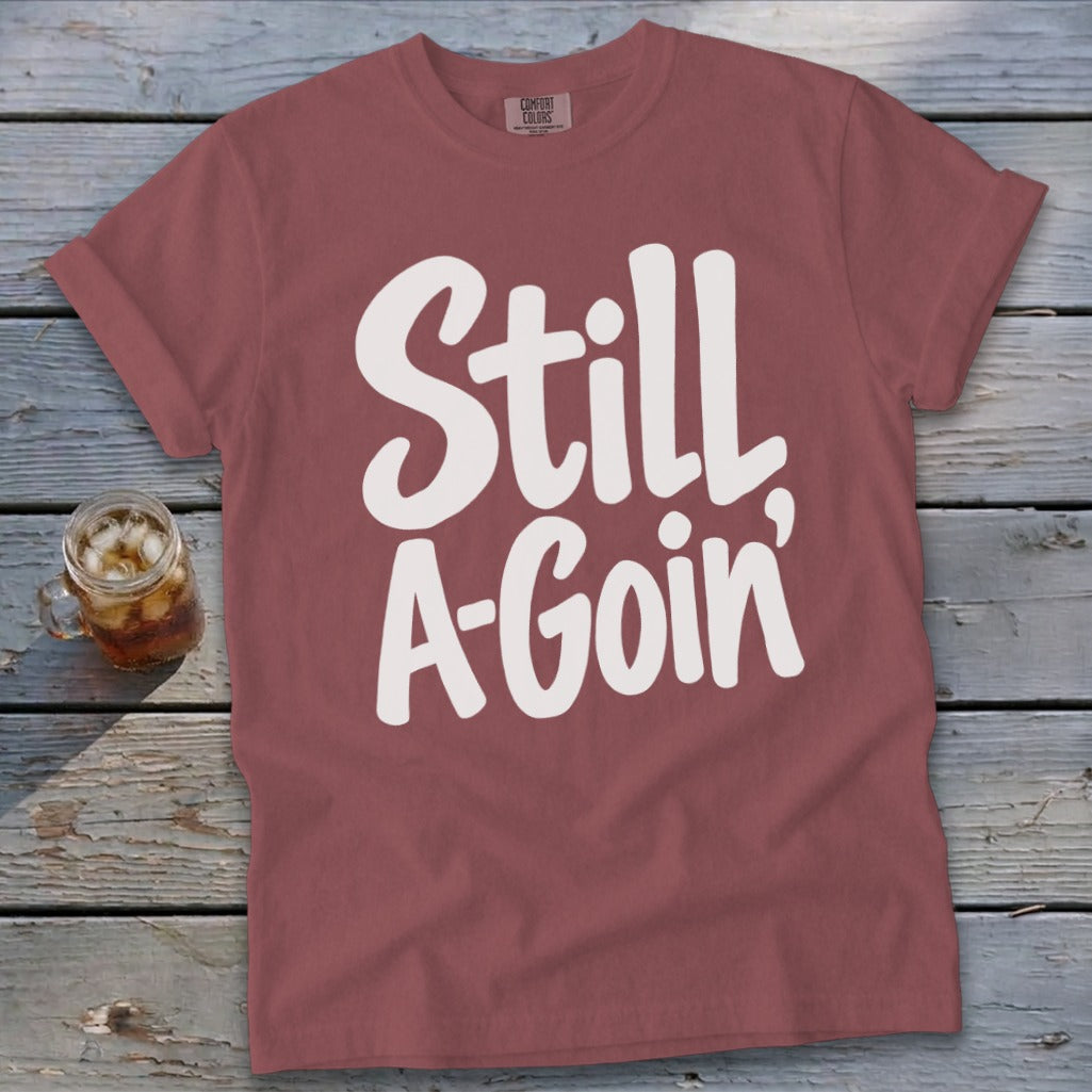 A dusty rose t-shirt featuring the white text Still A Goin lying flat on weathered wooden boards.