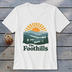 White v-neck t-shirt with graphic of sun setting over layered mountains and trees, reading "The Foothills".