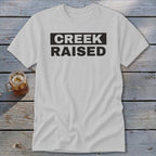 White t-shirt with black text that reads "CREEK RAISED" in a rectangular box.