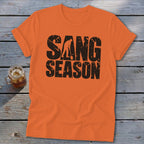 An orange t-shirt featuring distressed black text that reads SANG SEASON with a silhouette of a person digging.