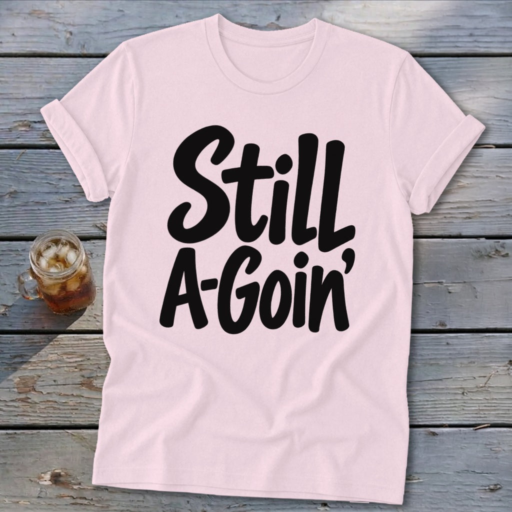 A light pink t-shirt with rolled sleeves features black text reading Still A-Goin’ on a wooden background.