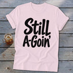 A light pink t-shirt with rolled sleeves features black text reading Still A-Goin’ on a wooden background.