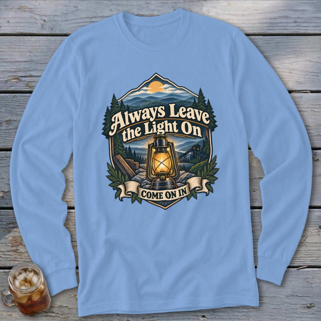 Light blue long sleeve shirt with a graphic featuring a lantern, mountains, and the text "Always Leave the Light On Come On In".