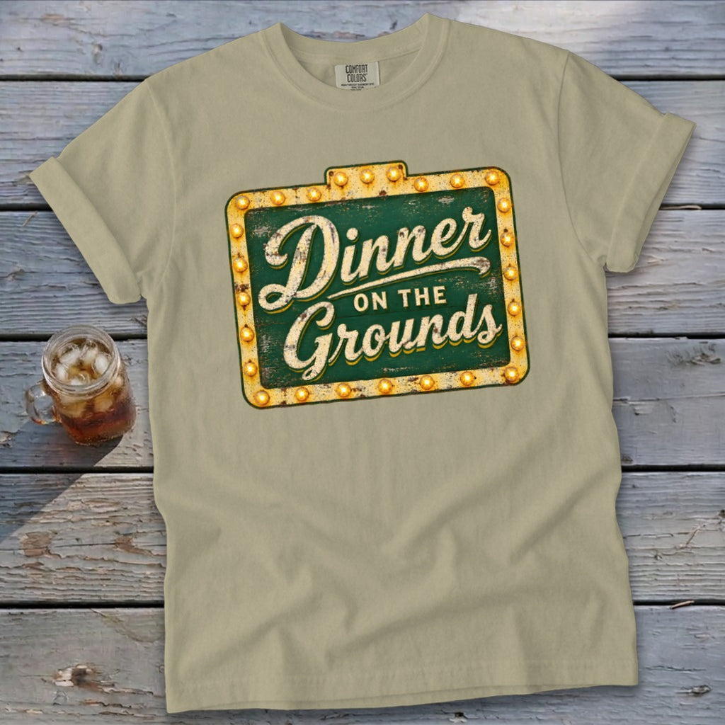 A light green crewneck t-shirt featuring a distressed green and yellow sign graphic that reads Dinner on the Grounds.