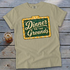 A light green crewneck t-shirt featuring a distressed green and yellow sign graphic that reads Dinner on the Grounds.