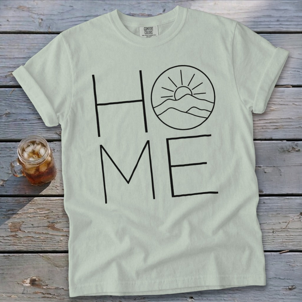 Light green t-shirt with black outline graphic spelling HOME, featuring a mountain sunrise inside the O.