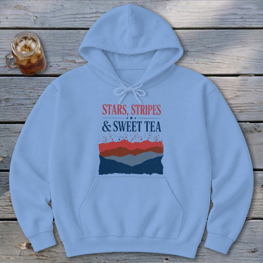 Light blue hooded sweatshirt with text and mountains graphic, laid flat on a rustic wooden surface next to a glass of iced tea.