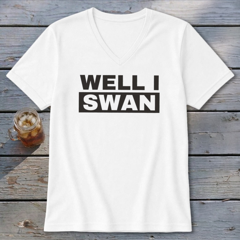 A white V-neck t-shirt with black text that reads WELL I SWAN lying on a wooden background with a glass of iced tea.