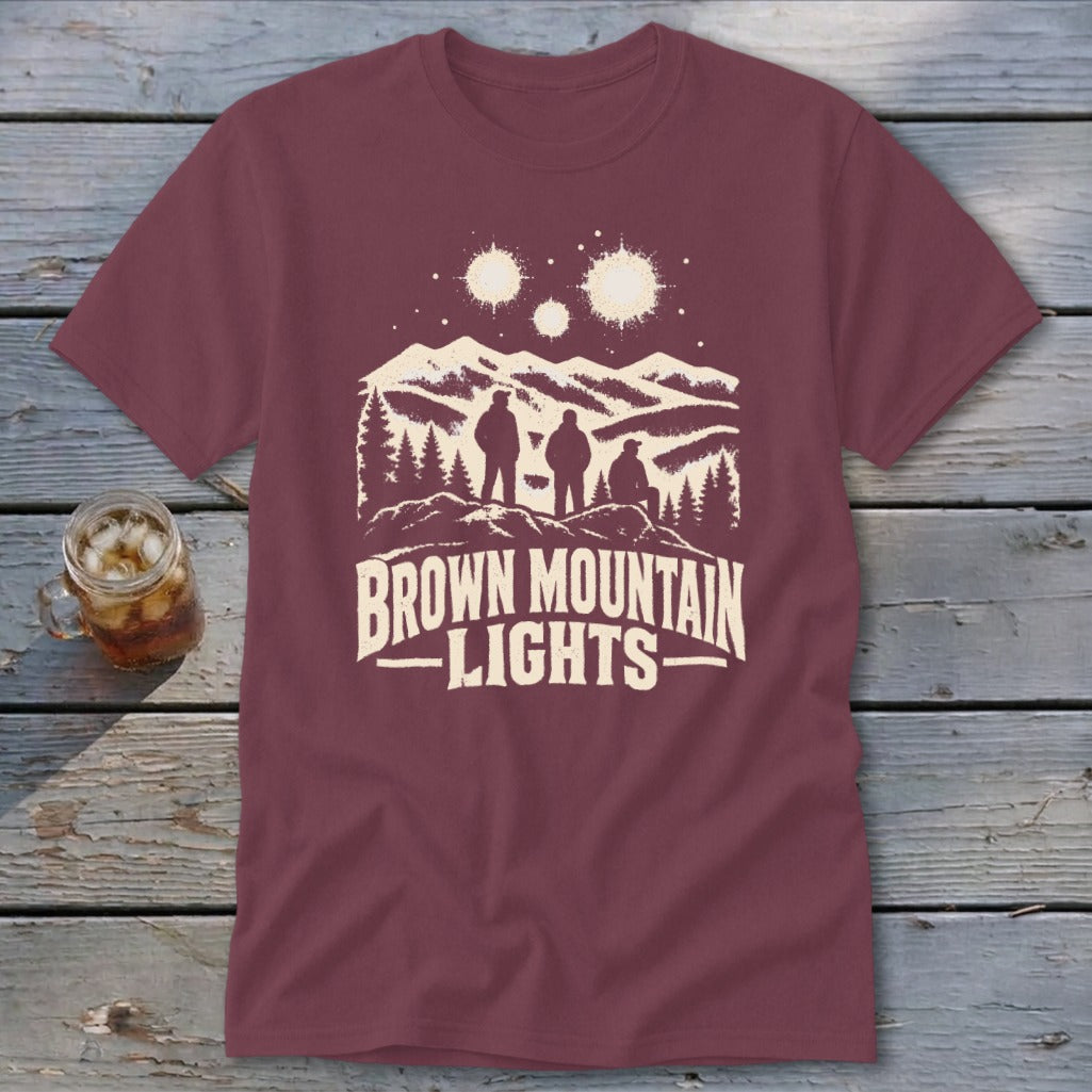 Maroon t-shirt featuring a graphic of three figures beneath glowing lights over mountains with text reading Brown Mountain Lights.