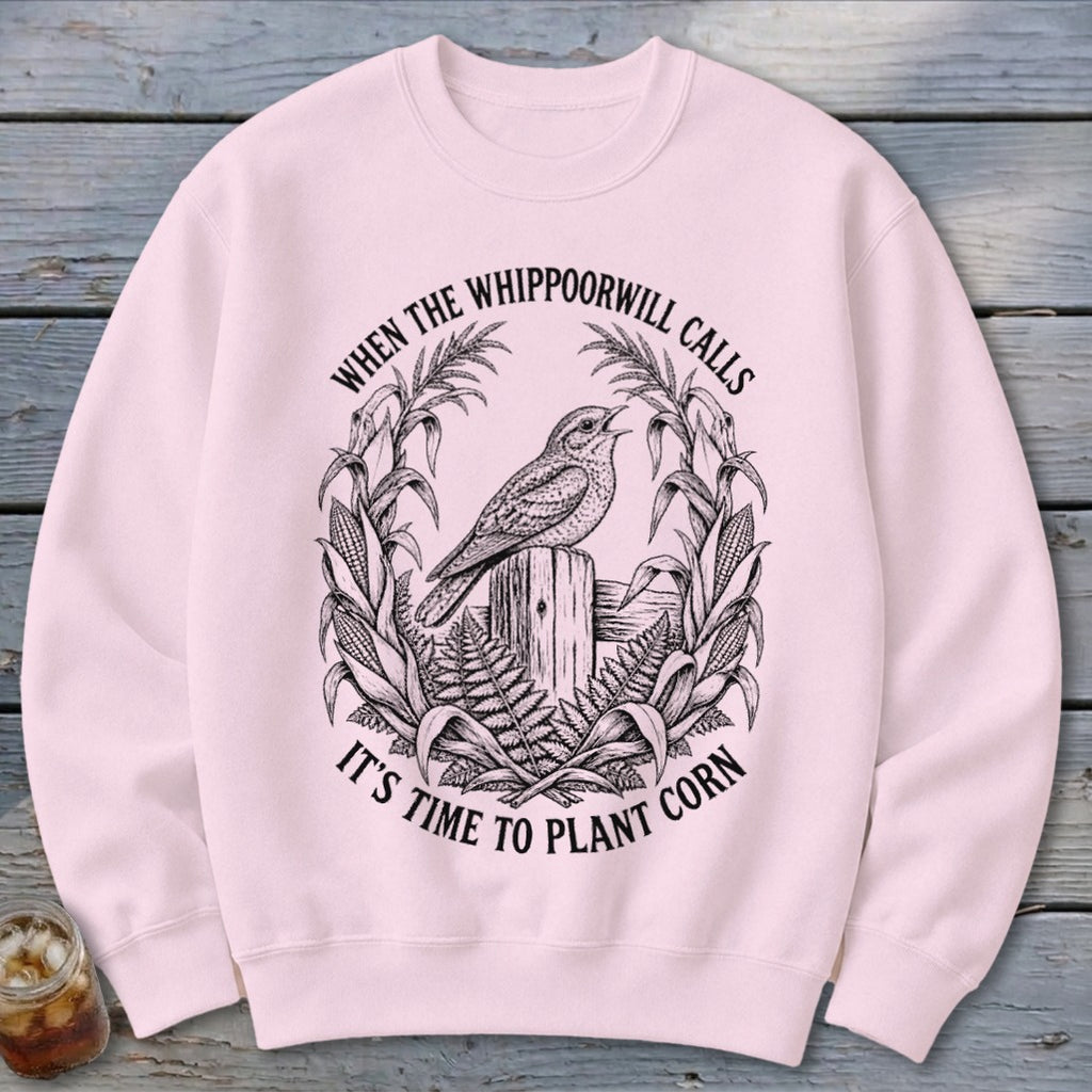 Light pink crewneck sweatshirt featuring a black vintage illustration of a bird on a fence post with the text "WHEN THE WHIPPOORWILL CALLS IT'S TIME TO PLANT CORN".