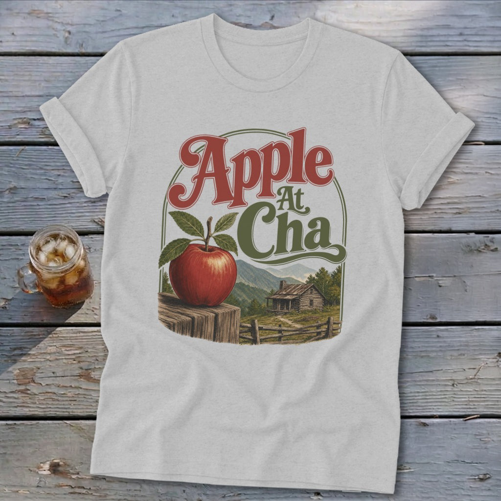 A light grey t-shirt featuring a graphic of an apple, a cabin, and mountains with the text Apple At Cha.