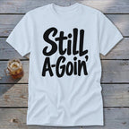 A white t-shirt with black cursive text reading "Still A-Goin'" lies flat on a weathered wooden surface next to a glass of iced tea.