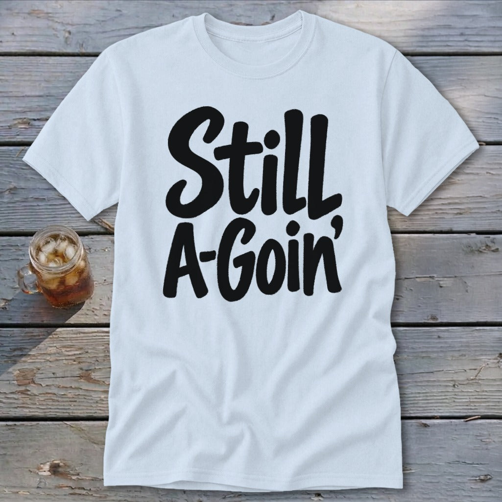 A white t-shirt with black cursive text reading "Still A-Goin'" lies flat on a weathered wooden surface next to a glass of iced tea.