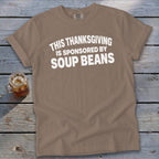 Brown t-shirt featuring the white text "THIS THANKSGIVING IS SPONSORED BY SOUP BEANS" laid on a wooden surface.