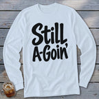 A white long-sleeved t-shirt with black cursive text reading Still A-Goin' on a wooden background.