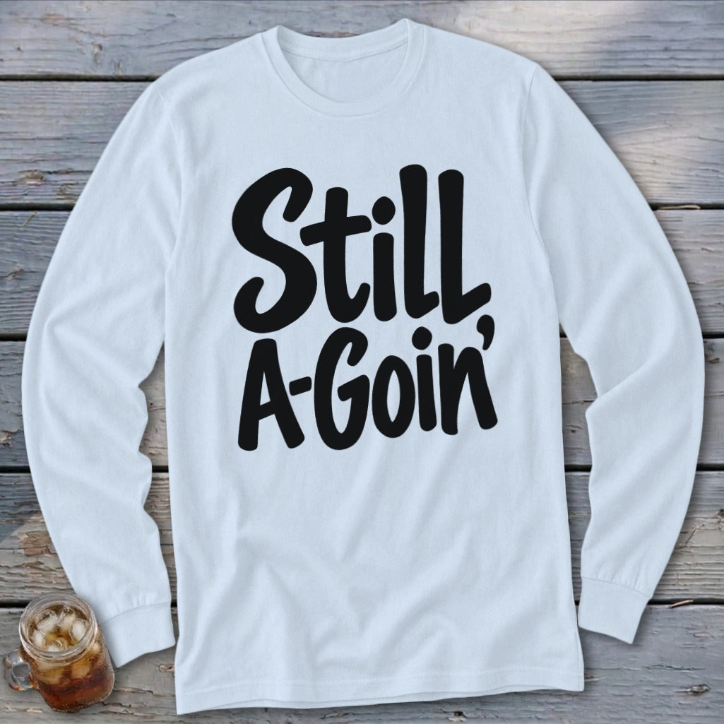 A white long-sleeved t-shirt with black cursive text reading Still A-Goin' on a wooden background.