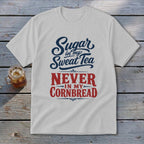 A light gray crewneck t-shirt featuring the blue and red graphic text "Sugar in my Sweet Tea NEVER IN MY CORNBREAD" laid flat on a rustic wooden surface next to a glass of iced tea.
