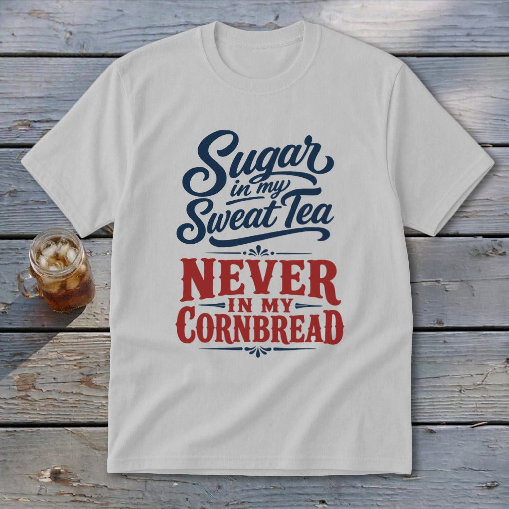 A light gray crewneck t-shirt featuring the blue and red graphic text "Sugar in my Sweet Tea NEVER IN MY CORNBREAD" laid flat on a rustic wooden surface next to a glass of iced tea.