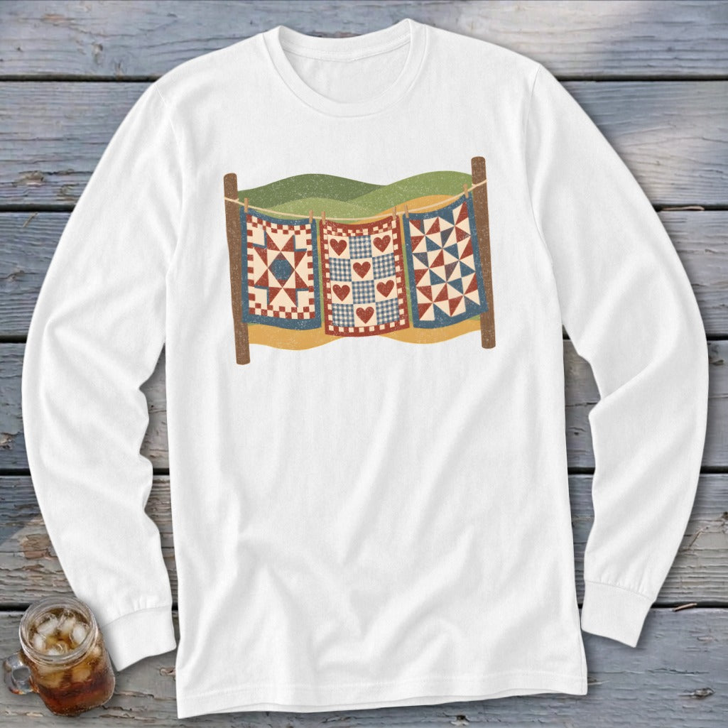 White long-sleeve t-shirt featuring a graphic of three vintage-style patchwork quilts hanging on a clothesline.