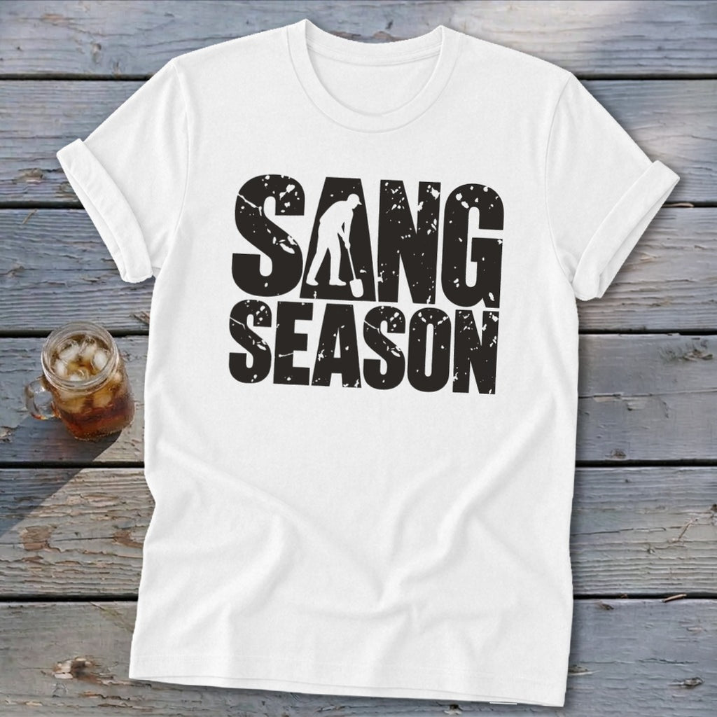 A white crew-neck t-shirt rests on weathered gray wood, featuring black distressed text that reads SANG SEASON with a silhouette of a person digging in the center.