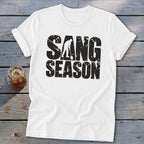 A white crew-neck t-shirt rests on weathered gray wood, featuring black distressed text that reads SANG SEASON with a silhouette of a person digging in the center.