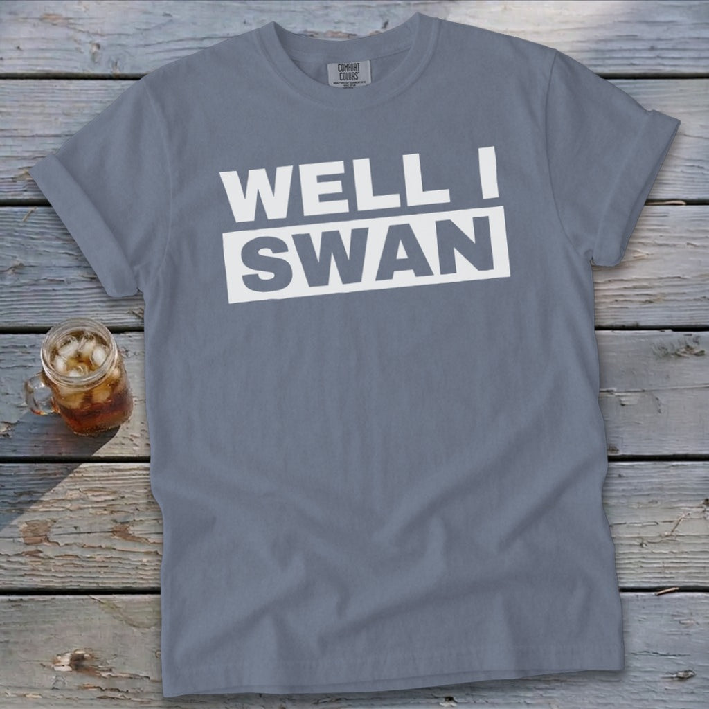 A slate blue t-shirt with the white text WELL I SWAN lying on a weathered wooden surface next to a glass of iced tea.