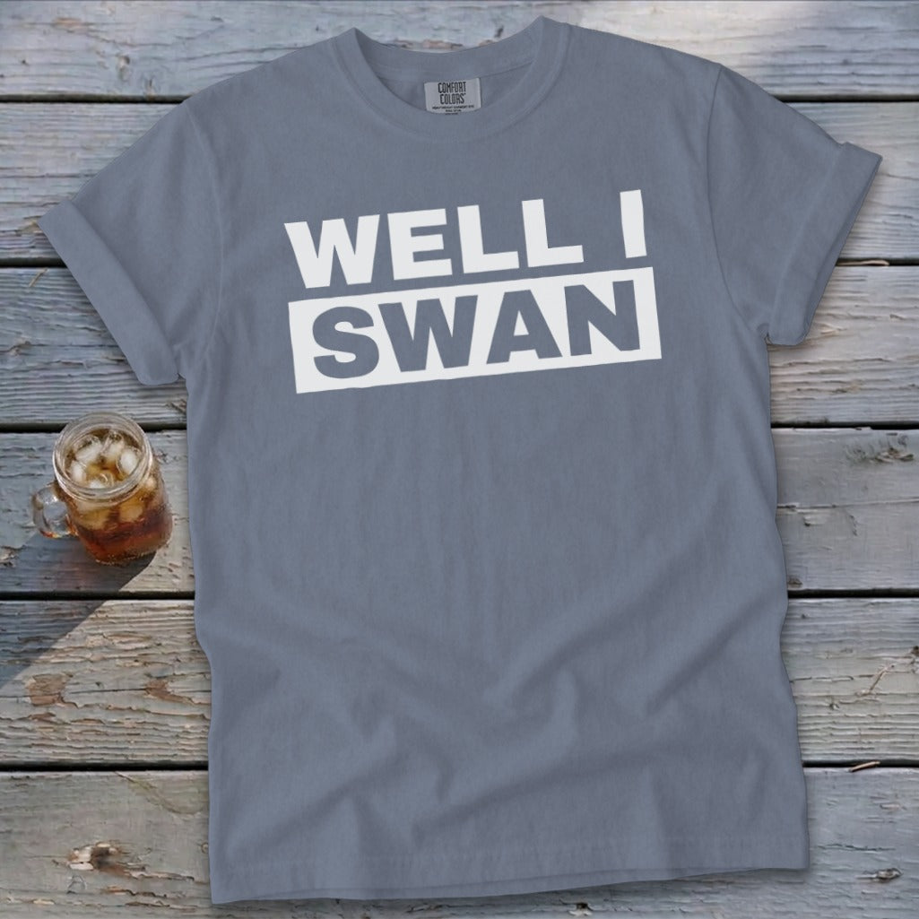 A slate blue t-shirt with the white text WELL I SWAN lying on a weathered wooden surface next to a glass of iced tea.