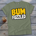 An olive green t-shirt with the text BUM FUZZLED on a wooden surface.