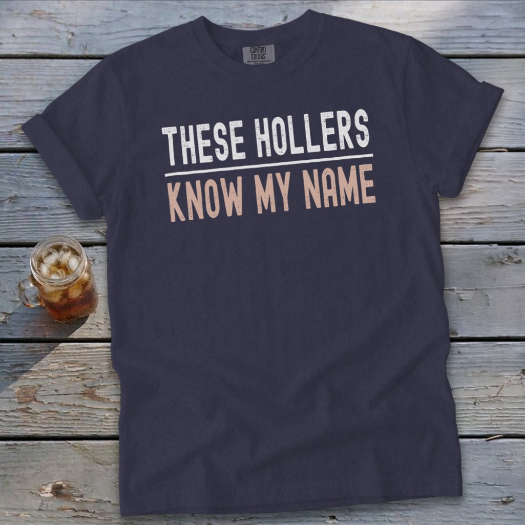 Navy blue t-shirt with white text "THESE HOLLERS" and rose text "KNOW MY NAME".