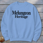 Light blue crewneck sweatshirt with the black text "Melungeon Heritage" centered on the chest, laid flat on a rustic wooden background.