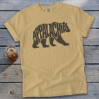 A tan t-shirt with a brown bear graphic containing the word Appalachia laid flat on a wooden surface.