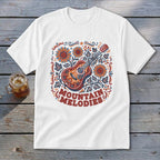 A white t-shirt featuring a colorful graphic of an acoustic guitar, sunflowers, leaves, and the text MOUNTAIN MELODIES.