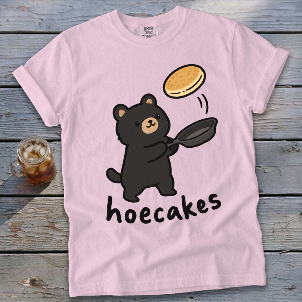 A light pink t-shirt featuring a cartoon black bear flipping a pancake from a frying pan above the text hoecakes.