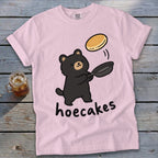 A light pink t-shirt featuring a cartoon black bear flipping a pancake from a frying pan above the text hoecakes.