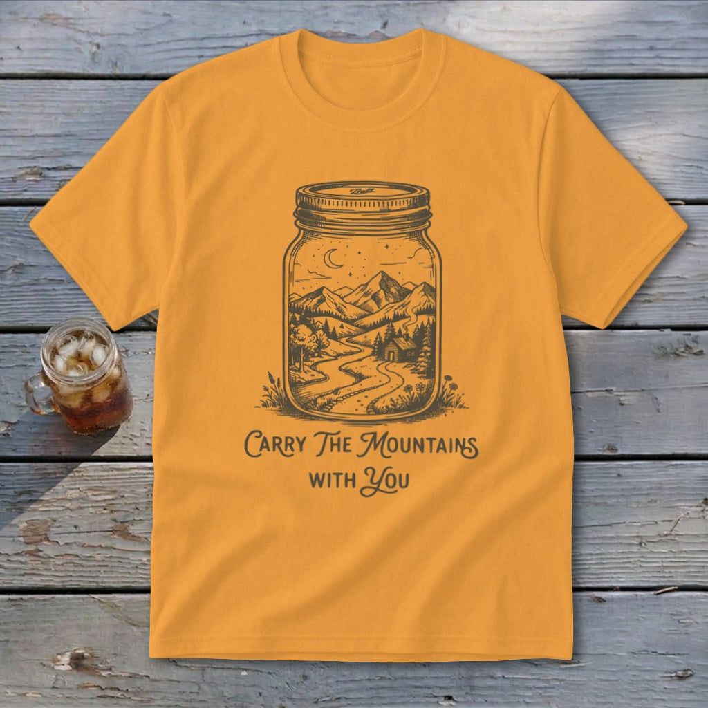A mustard yellow t-shirt features a black illustration of a mountain landscape inside a mason jar, with the text "Carry The Mountains With You."