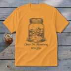 A mustard yellow t-shirt features a black illustration of a mountain landscape inside a mason jar, with the text "Carry The Mountains With You."